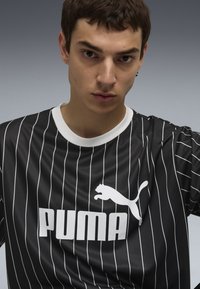 Cămașă sport neagră, cu dungi, având accente albe. Dispune de un guler rotund și un logo alb Puma proeminent pe piept. Fabricată dintr-un material neted.