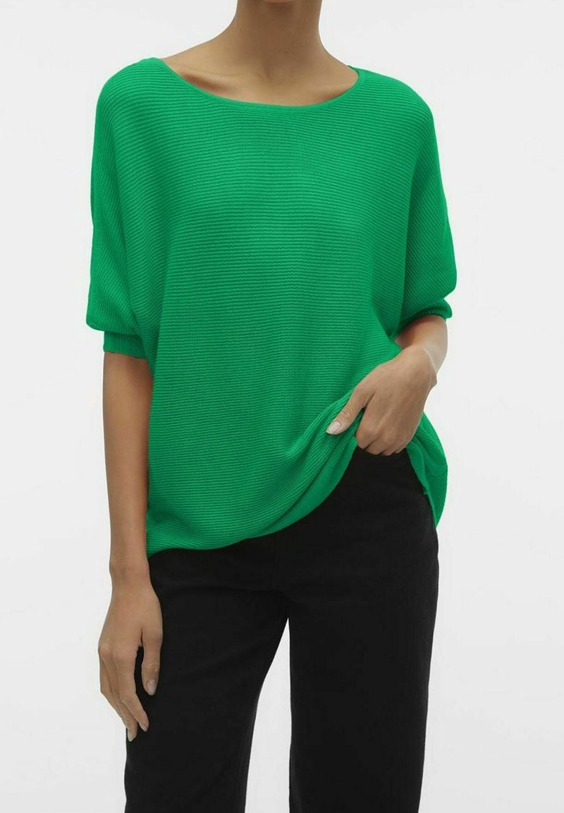 Personne portant un pull léger vert vif à manches courtes côtelé et un pantalon noir, une main dans la poche et l'autre détendue.