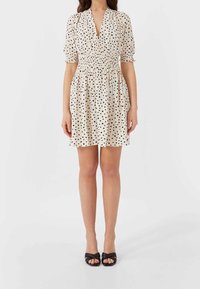 Helles beige Kleid mit schwarzen Punkten, kurzen Puffärmel, V-Ausschnitt und gerüschtem Taillenbund. Getragen mit schwarzen offenen Sandalen mit Absatz.