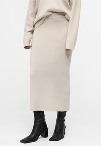 Jupe maxi en maille beige avec une taille ajustée, une texture lisse et une fente latérale, associée à des bottines en cuir noir avec bouts carrés.