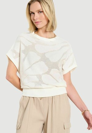 Donna bionda che indossa un top a maniche corte bianco lavorato a maglia con motivi e pantaloni beige a vita alta, in piedi davanti a uno sfondo chiaro e semplice.