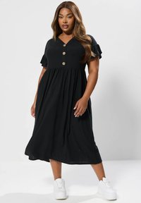 Czarna, prążkowana sukienka midi z krótkimi, falbaniastymi rękawami i dekoltem w kształcie litery V; posiada trzy guziki z przodu i rozkloszowaną spódnicę. Stylizowana z białymi trampkami.