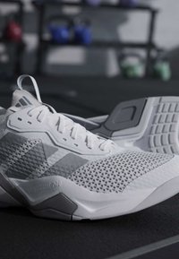 Κοντινό πλάνο λευκών αθλητικών παπουτσιών Adidas με υφασμένο διχτυωτό σχέδιο και λαστιχένιες σόλες σε δάπεδο γυμναστηρίου με θολωμένα βάρη στο φόντο.