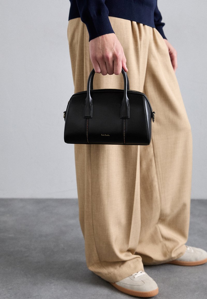 Black leather handbag with structured design, dual handles, subtle stitching details, and a gold logo. Paired with beige wide-leg trousers.