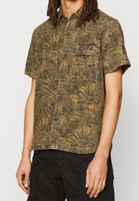 Olijfgroen shirt met korte mouwen, voorzien van een loofpatroon, twee borstzakken, blauwe knopen en een oranje logo-detail op de mouw.