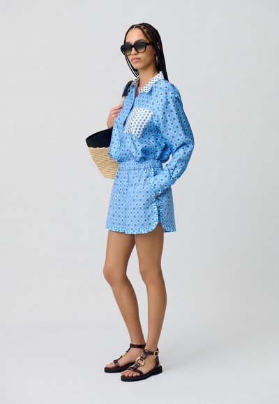 Vestido corto de estampado azul con cuello y cintura elástica. Llevado con sandalias planas marrones y un bolso de rafia. Grandes gafas de sol completan el look.