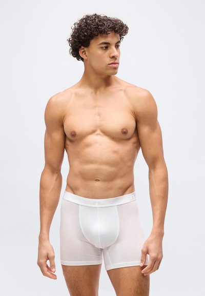 Μυώδης νεαρός με σγουρά μαλλιά, ντυμένος με λευκά ανδρικά εσώρουχα τύπου μπόξερ, στέκεται και κοιτάει προς τα πλάγια μπροστά σε απλό φωτεινό φόντο.