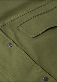 Gros plan sur un tissu vert olive avec une poche à rabat cousue et deux boutons-pression métalliques sur un vêtement.