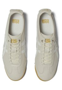 Béžové sportovní boty vyrobené z kůže s bílými pruhy, šněrovací design a strukturovanou stélkou. Na jazyku se nachází zlaté logo.