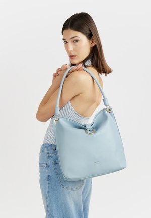 Donna con capelli castani che indossa un top senza maniche e jeans tiene una grande borsa in pelle azzurra sulla spalla davanti a uno sfondo bianco.