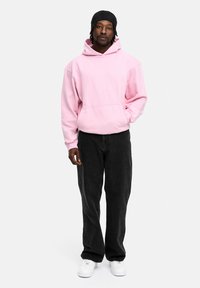 Light pink hoodie with a kangaroo pocket, paired with black wide-leg jeans and white sneakers. The hoodie features a drawstring hood.
