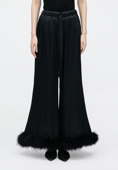 Pantalon noir à jambes larges avec finition satinée ; taille élastique, ceinture assortie et bordure en fausse fourrure à l'ourlet. Porté avec des chaussures noires pointues.