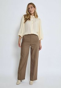 Cremefarbige Bluse mit Knopfleiste und puffärmel, kombiniert mit braunen High-Waist-Hosen und beigen Absätzen. Schlichtes, strukturiertes Design.