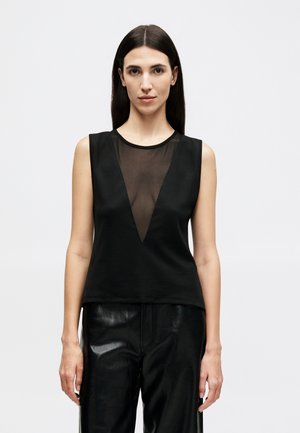 Moteris su ilgais tamsiais plaukais dėvi juodą rankovių neturinčią palaidinę su permatomu V formos įsiuvimu ir blizgusiomis juodomis kelnėmis, stovinti priešais vienspalvį foną.