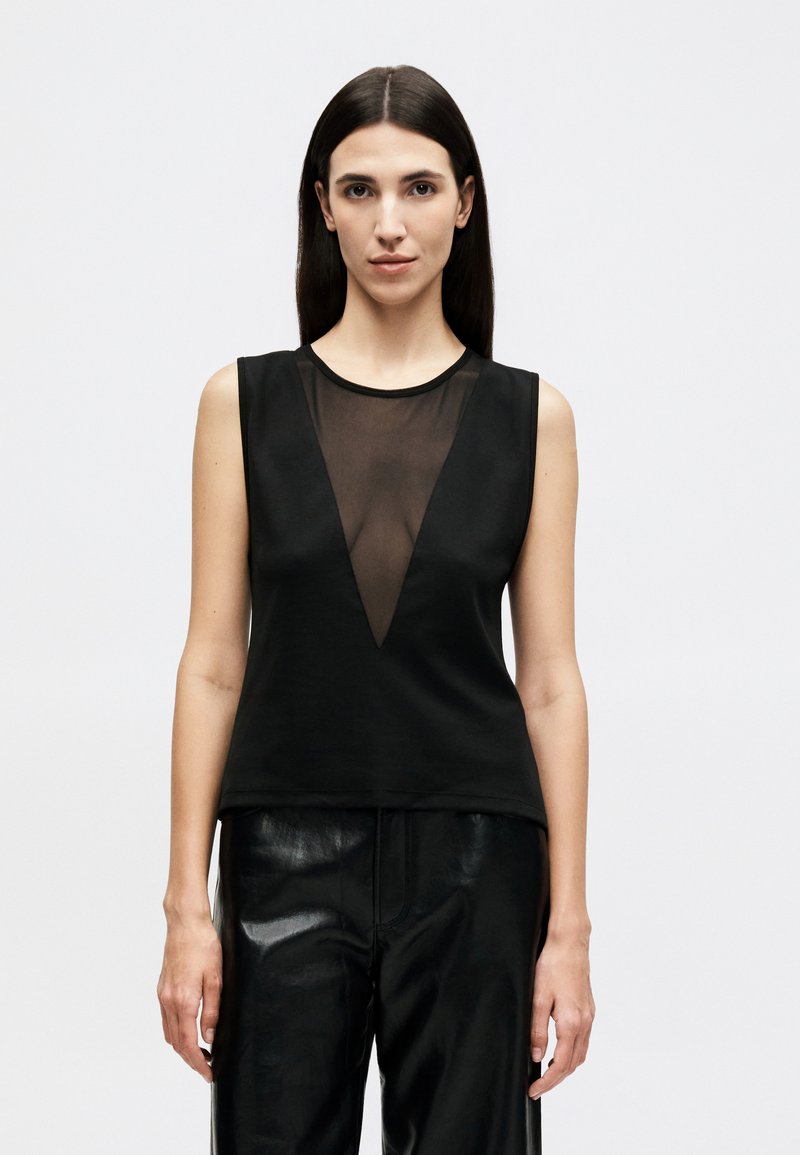 Kvinde med langt mørkt hår iført sort, ærmeløs top med gennemsigtigt V-formet panel og sorte skinnende bukser, står mod en ensfarvet baggrund.