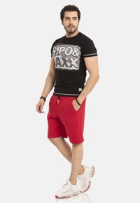 Zwart t-shirt met witte grafische tekst, gecombineerd met rode shorts met zijzakken, en witte sneakers met rode accenten.