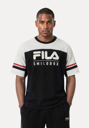 Mand iført sort og hvid FILA SMILODOX t-shirt med røde og sorte striber på ærmerne, stående med hænderne i lommerne mod en ensfarvet baggrund.