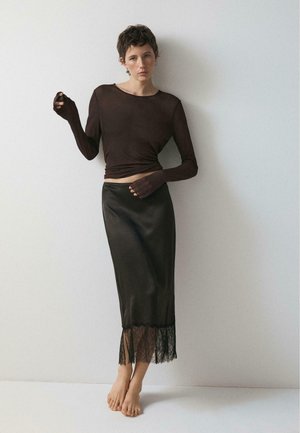 Donna con capelli corti che indossa un top nero aderente a maniche lunghe e una gonna midi di raso nera con orlo in pizzo, in piedi scalza contro un muro semplice.