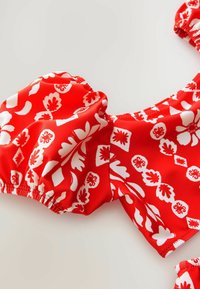 Czerwony biustonosz bikini w kwiaty z białymi wzorami liści i kwiatów, elastycznymi falbaniastymi brzegami oraz gładką, błyszczącą teksturą materiału.