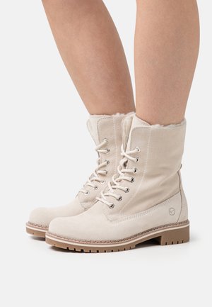 Botas de tobillo de ante beige con un suave forro de piel, que cuentan con ojales metálicos y cordones redondos, además de una suela de goma resistente para mayor tracción.