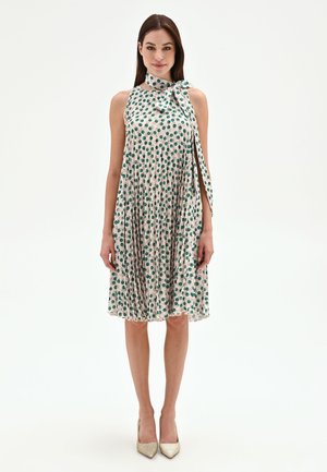 Donna che indossa un vestito beige senza maniche fino al ginocchio con pois verdi e scollo con fiocco, abbinato a décolleté beige.