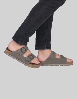 Personne portant des sandales grises à boucle réglable avec semelles en liège et un jean noir sur un fond gris uni.