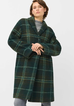 Woman wearing a dark green oversized plaid coat with large collar and button, standing against a white background.