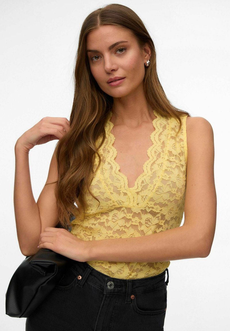 Jeune femme aux longs cheveux bruns, portant un haut sans manches en dentelle jaune et un pantalon noir, tenant une veste en cuir noire.