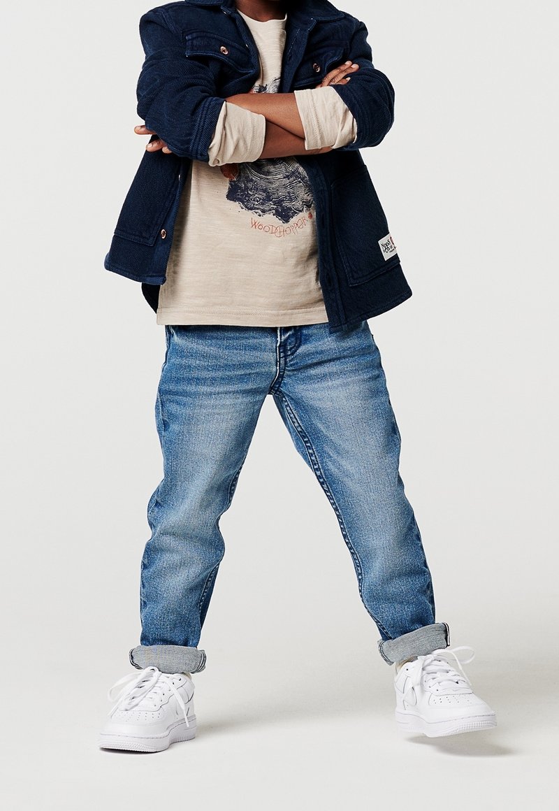 Enfant debout, les bras croisés, portant une veste bleu marine, une chemise beige imprimée, un jean bleu retroussé et des baskets blanches.