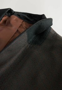 Dunkel gemusterte Blazer-Innenausstattung mit einem glatten braunen Futter; verfügt über einen strukturierten schwarzen Stoff und eine kleine Stoffschleife in der Nähe des Kragens.