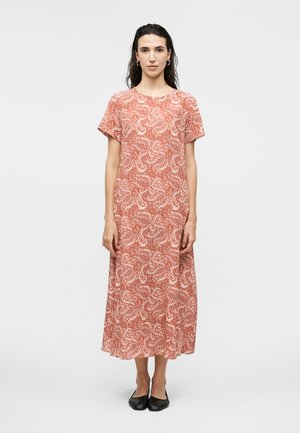 Γυναίκα στέκεται με το πρόσωπο προς τα εμπρός, φορώντας ένα φορεματάκι μέτριου μήκους με κοντά μανίκια, πορτοκαλί και λευκό με μοτίβο, και μαύρες μπαλαρίνες, σε απλό φόντο.