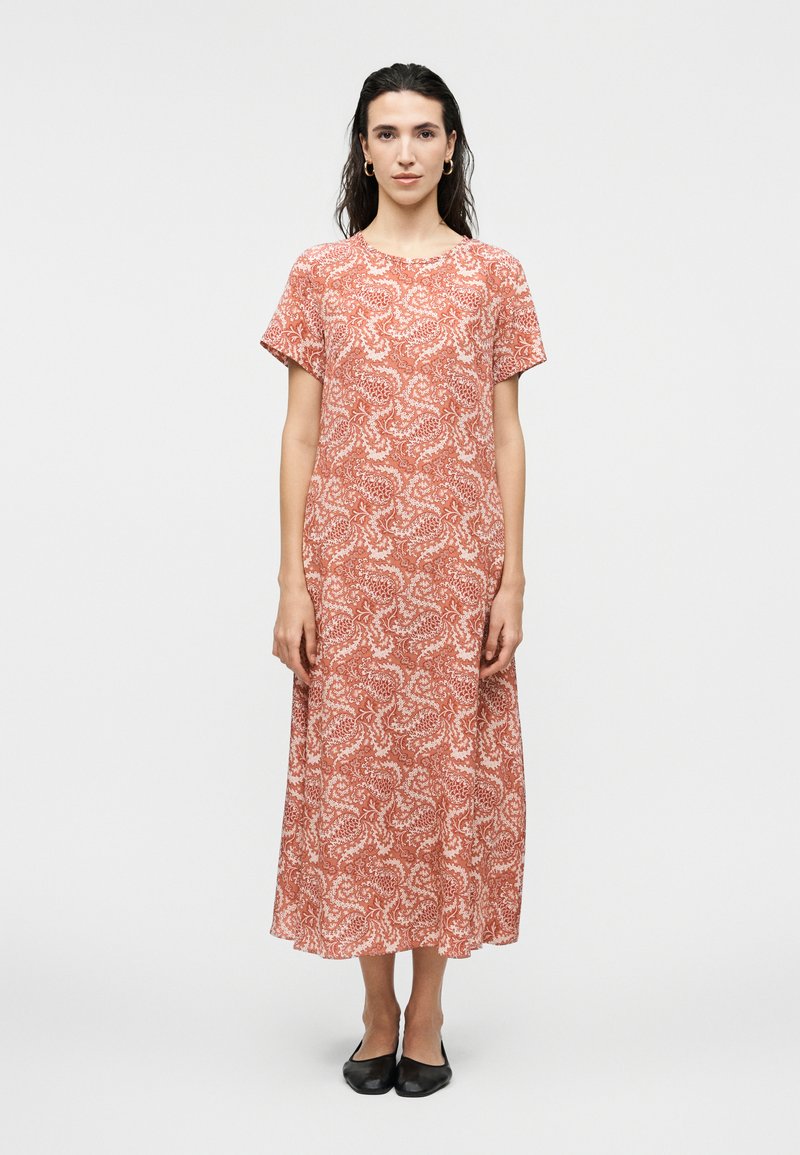 Γυναίκα στέκεται με το πρόσωπο προς τα εμπρός, φορώντας ένα φορεματάκι μέτριου μήκους με κοντά μανίκια, πορτοκαλί και λευκό με μοτίβο, και μαύρες μπαλαρίνες, σε απλό φόντο.