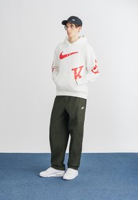 Bela kapuca s rdečim Nike logotipom spredaj in na rokavih, olivno zeleni cargo hlače in bele tenisice. Črna kapa z detajli logotipa.
