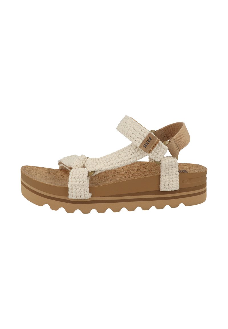 Beige woven sandals with a cork footbed, adjustable straps, and a brown rubber sole featuring a wavy tread pattern for traction.