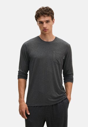 Giovane uomo con capelli corti e ricci che indossa una camicia a maniche lunghe grigio scuro con taschino sul petto e pantaloni grigio scuro abbinati, in piedi davanti a uno sfondo chiaro.