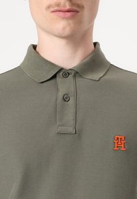 Olivově zelené polo tričko s texturovaným pleteným materiálem, se dvěma černými knoflíky na límci a malým oranžovým vyšitým logem na prsou.