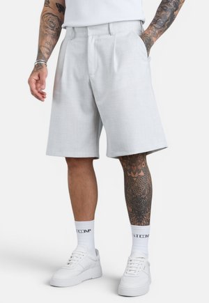 Mann trägt hellgraue knielange Shorts, weiße Socken und weiße Turnschuhe, mit tätowierten Armen und einem sichtbaren tätowierten Bein.