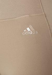 Leggings atletici beige realizzati in tessuto elasticizzato con una vita a coste, dotati di un logo Adidas riflettente e dettagli lisci e senza cuciture.
