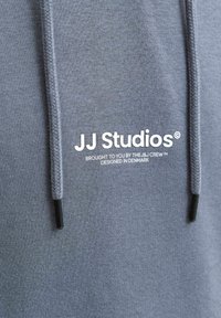 Gros plan d'un sweat à capuche en tissu gris avec le texte blanc "JJ Studios" et des cordons avec embouts noirs.