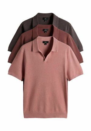 Mężczyzna o kręconych włosach, ubrany w polo z krótkim rękawem w kolorze rdzawym, czarny pasek, czarne spodnie i czarne mokasyny, stoi ze złożonymi rękami w kieszeniach.