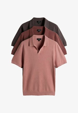 Man met krullend haar, gekleed in een roestkleurig poloshirt met korte mouwen, zwarte riem, zwarte broek en zwarte loafers, staand met handen in de zakken.