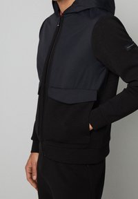 Sudadera con cremallera negra, con un panel oscuro en contraste, bolsillos frontales y capucha. Fabricada con un tejido suave y de textura lisa, con un diseño ajustado.