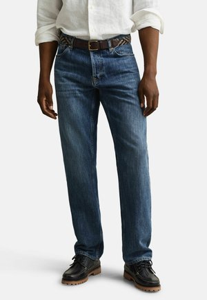 Mann trägt blaue Jeans mit geradem Bein, dunklem Ledergürtel, weißem hochgekrempeltem Hemd und schwarzen Schnürschuhen mit braunen Sohlen, steht vor weißem Hintergrund.