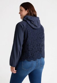 Granatowa kurtka z kapturem z koronkowym panelem na plecach, teksturowanym przodem i długimi rękawami, w połączeniu z niebieskimi dżinsami.