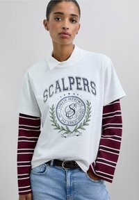 Camisa polo blanca con un logo y un diseño de hoja verde, superpuesta sobre una camisa de manga larga a rayas burdeos. Llevada con jeans azules y un cinturón negro.