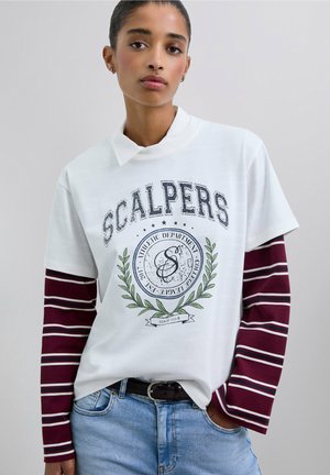 Camisa polo blanca con un logo y un diseño de hoja verde, superpuesta sobre una camisa de manga larga a rayas burdeos. Llevada con jeans azules y un cinturón negro.