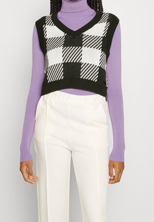 Vrouw draagt een zwart-wit geruite cropped gilet over een lavendelkleurige coltrui met witte broek met hoge taille.
