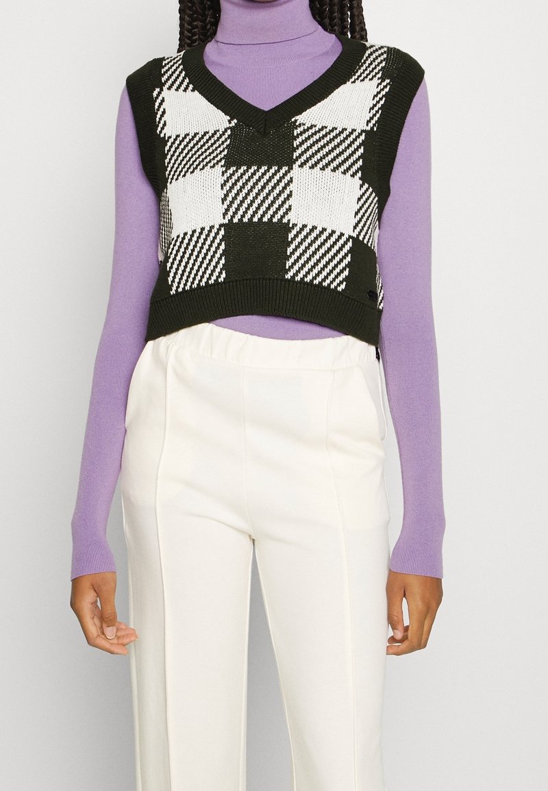 Femme portant un gilet court à carreaux noir et blanc sur un col roulé lavande avec un pantalon blanc taille haute.