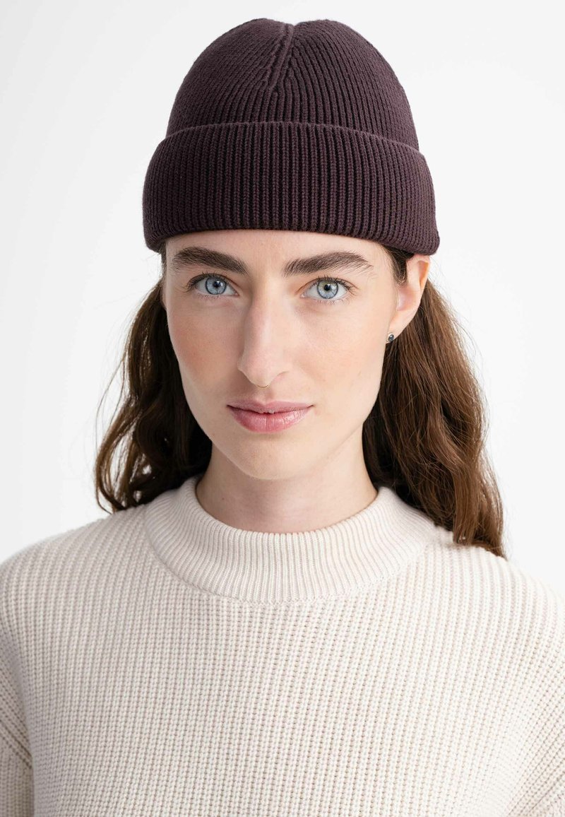 Dark purple knitted beanie with a ribbed texture and a seam at the top, worn with a light beige, textured sweater.