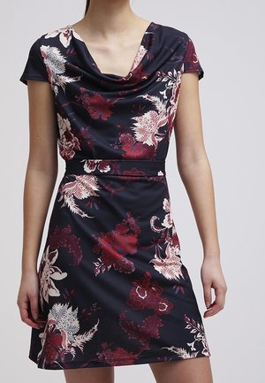 Femme portant une robe courte à manches courtes, de couleur marine avec des motifs floraux rouges et blancs, et un décolleté drapé, sur un fond uni.